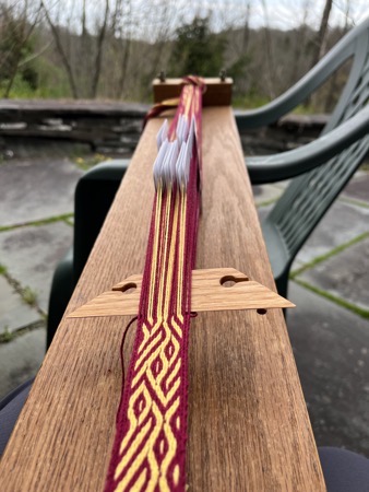 Wooden loom with red and gold band in progress.